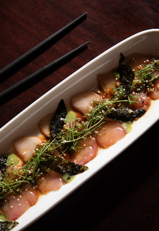 Delicate raw fish slices garnished with green herbs and sesame seeds on a white rectangular plate with chopsticks beside.