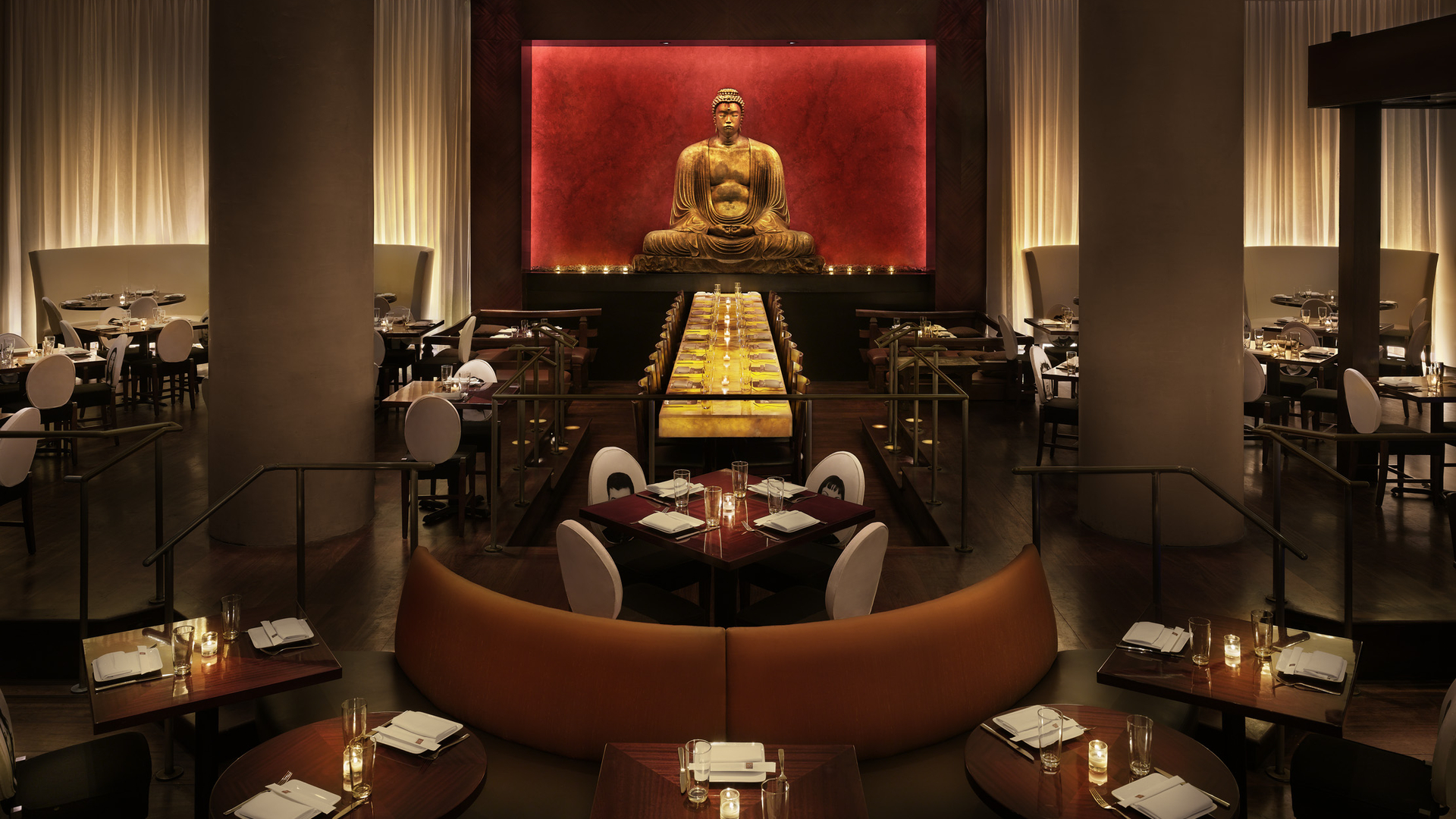 Modern restaurant with long table, Buddha statue in background, warm lighting, orange booths, multiple dining tables.