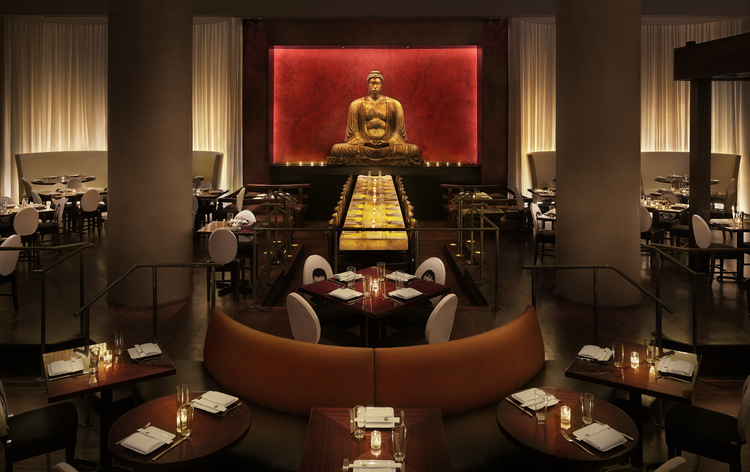 Modern restaurant interior with long table, Buddha statue, red backdrop, multiple round tables, warm lighting, and orange seating.
