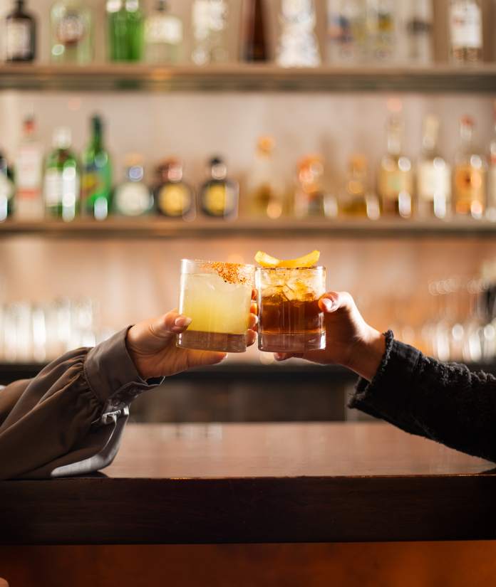 Two hands holding colorful cocktails at a bar with blurred bottles in the background.