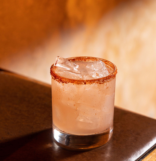 Cocktail with pinkish liquid, ice cubes, and a cinnamon rim, served in a short glass on a wooden bar.