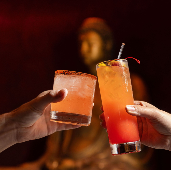 Two hands holding colorful drinks, one with a cherry garnish, in front of an ornate, gold Buddha statue.