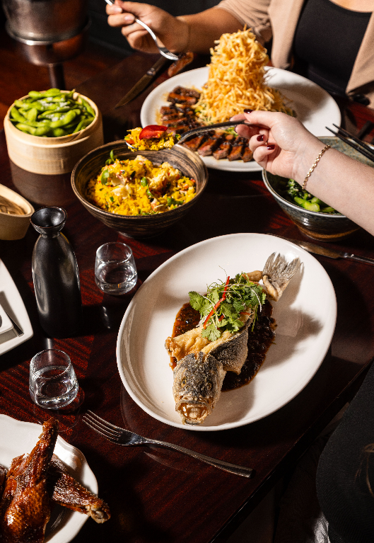 Overhead view of a shared dinner table with multiple dishes including a whole roasted fish garnished with microgreens, a grilled meat dish, yellow fried rice, edamame, a crispy potato nest, and a roasted chicken, with diners' hands reaching across the table.