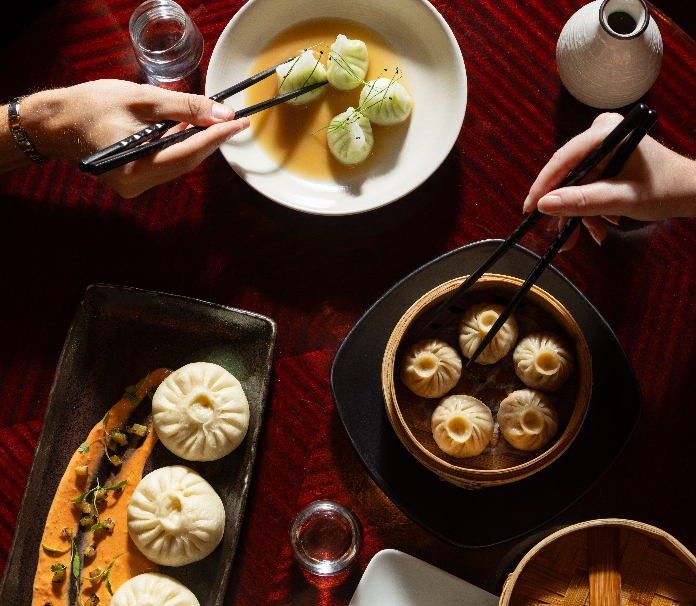Overhead view of two people using chopsticks to pick dumplings from a bamboo steamer basket and a white bowl, alongside a rectangular dish of steamed buns on a dark red textured table.
