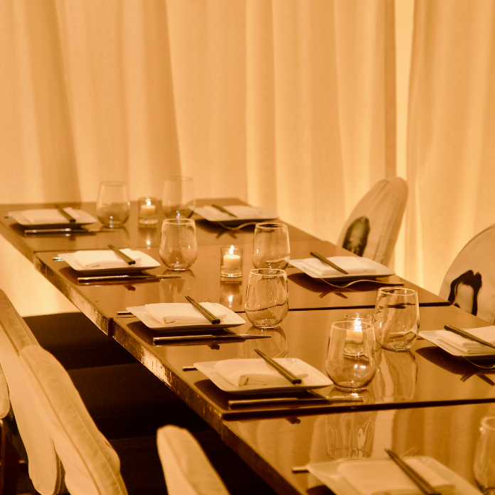 A warmly lit private dining room with a long dark table set for a group, featuring chopsticks, white place settings, wine glasses, and small candles against floor-to-ceiling ivory drapes.