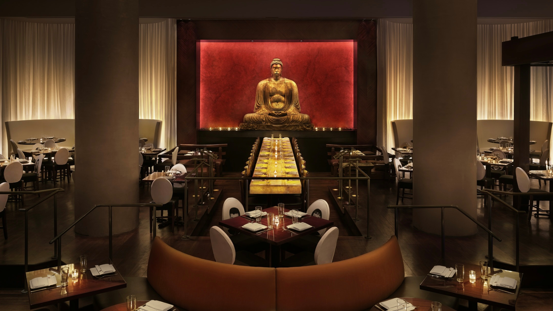 Modern restaurant with long table, Buddha statue in background, warm lighting, orange booths, multiple dining tables.