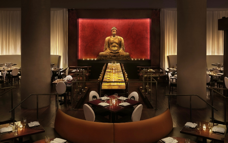 Modern restaurant with long table, Buddha statue in background, warm lighting, orange booths, multiple dining tables.