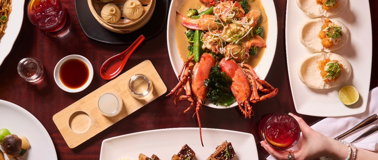 Overhead view of an elaborate Asian dining spread featuring a whole lobster, a seafood noodle dish, steamed dumplings, open-faced buns with toppings, a sake flight, dipping sauces, and a cocktail held by a hand at the edge of the table.